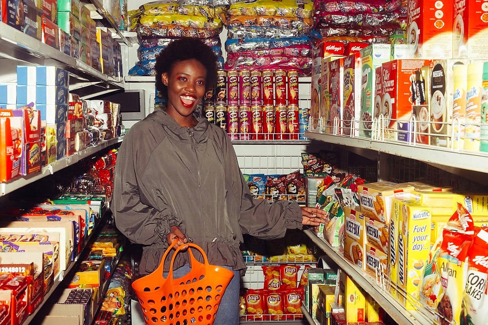 Free Happy African American lady in casual outfit with shopping basket in hand choosing food from products in small supermarket among colorful boxes on shelves and smiling with mouth opened Stock Photo
